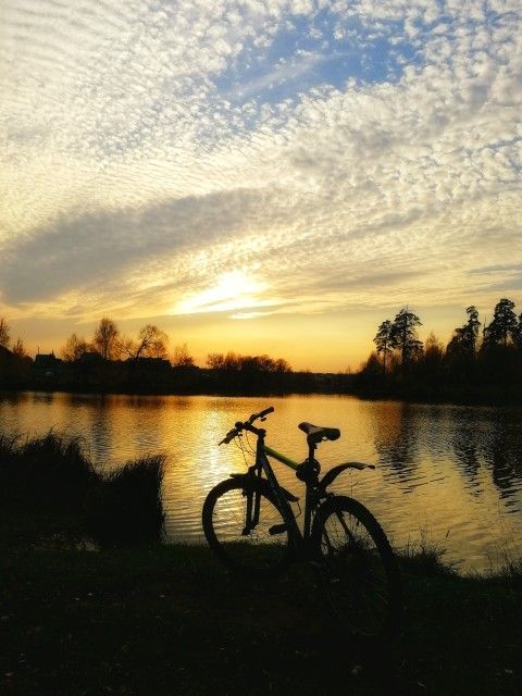 Silhouetted mountain bike on grass beside a calm lake at sunset with golden reflections on the water and a cloudy sky
